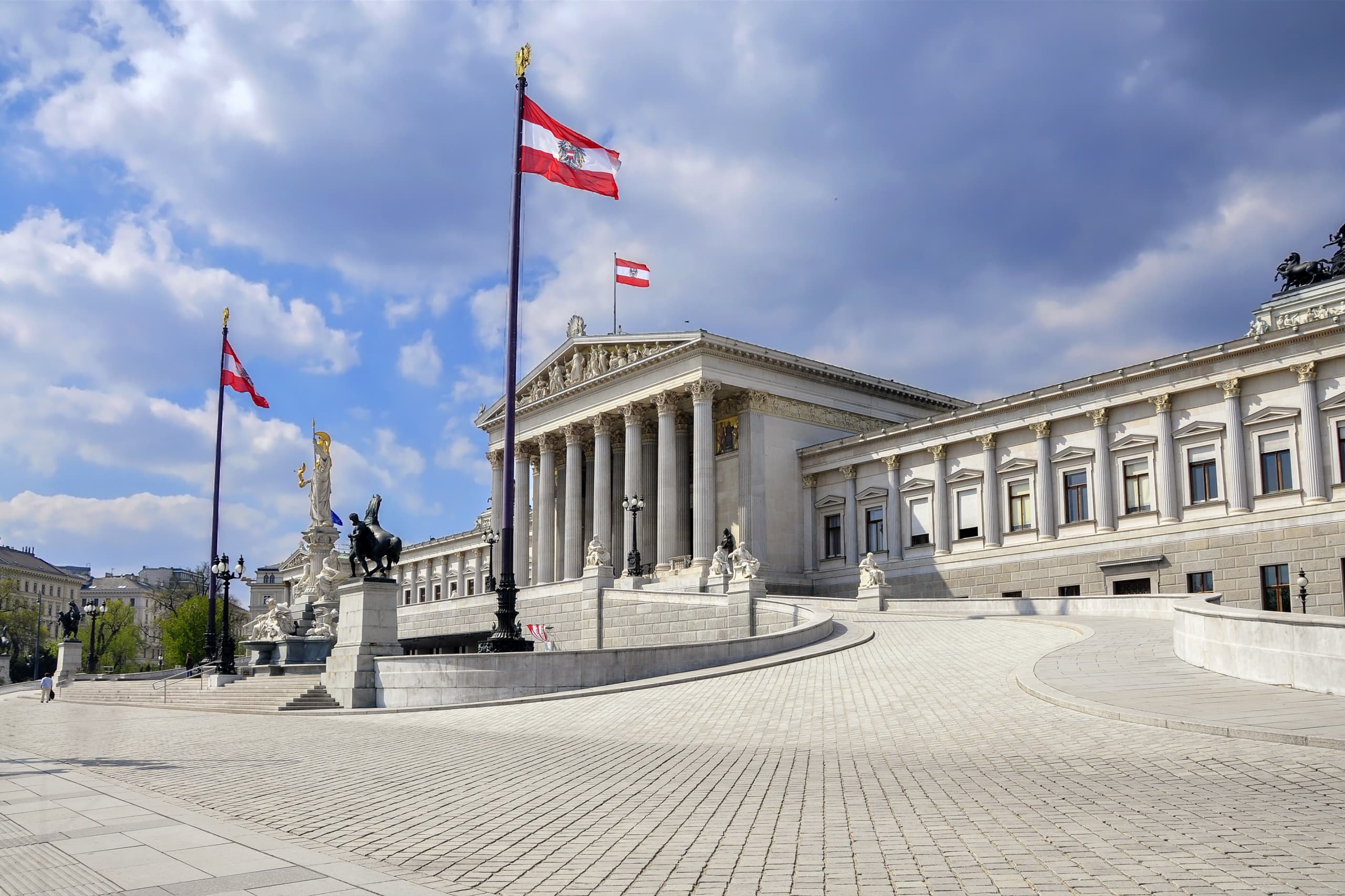 The Austrian Parliament Building in Vienna radiates energy with its neoclassical architecture, grand columns, statues, and red-and-white flags flying proudly against a cloudy blue sky.