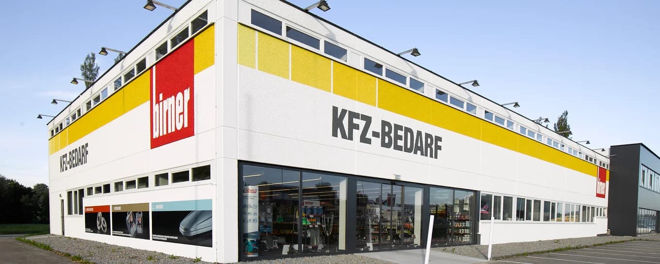 A large auto parts store with KFZ-BEDARF on the white and yellow facade, big glass windows showing products inside, and a clear sky above—now enhanced with LKUF AI Solution Data Processing for efficient inventory management.
