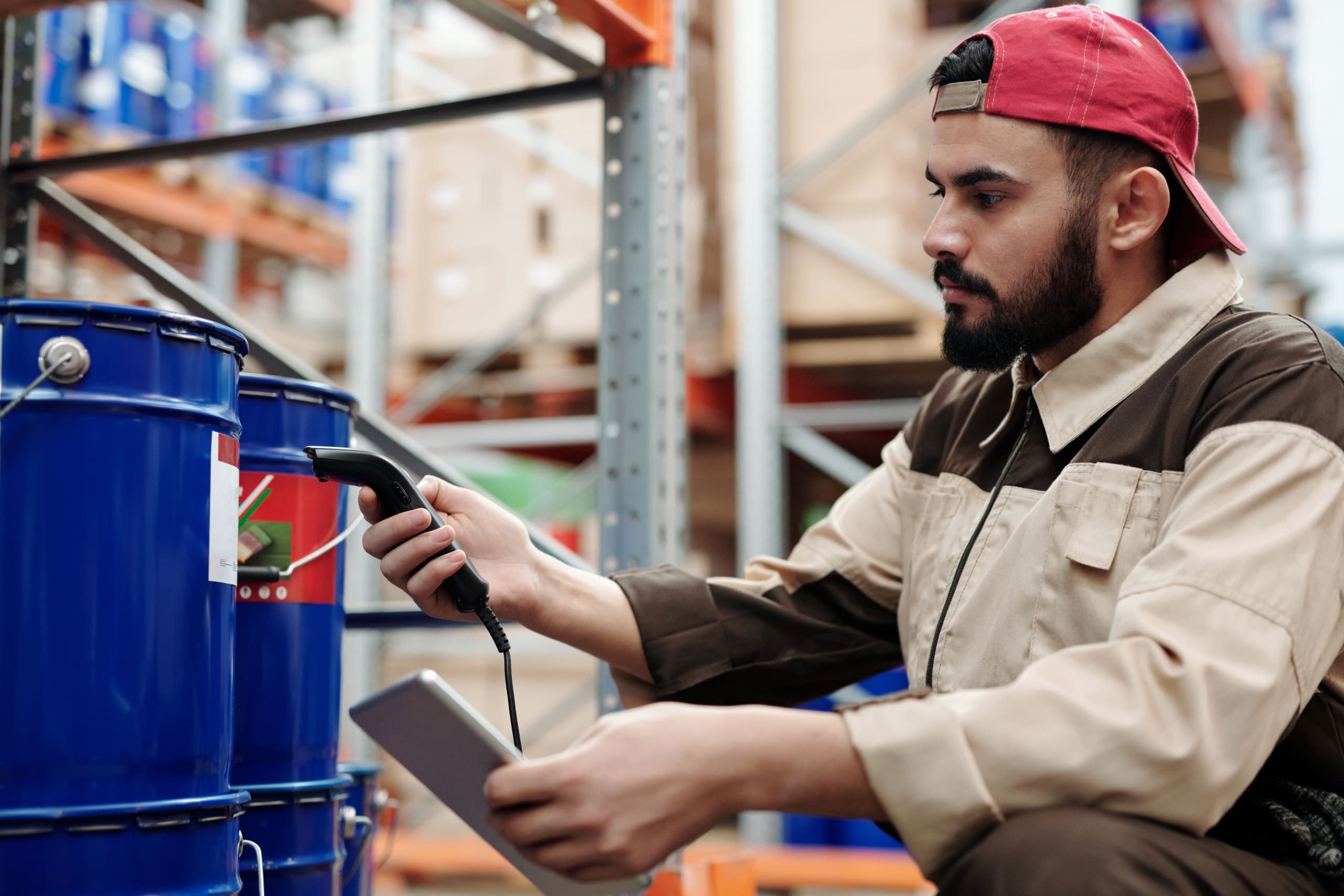 A warehouse worker in a tan and brown uniform scans a blue barrel with a handheld barcode scanner while holding a tablet loaded with AI-ready product data. He is wearing a red backward cap and appears focused on his task. Shelves are visible in the background, highlighting the efficiency brought by OpenAI checkout systems.